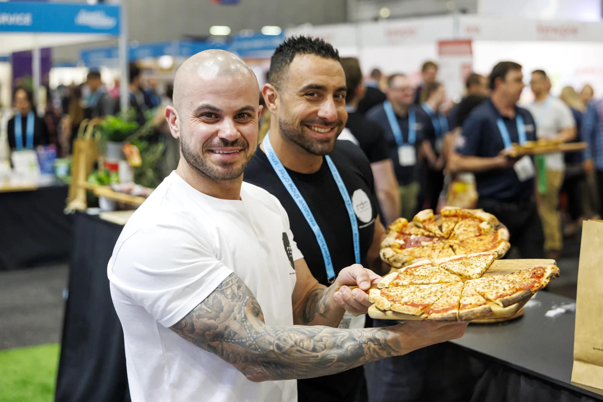 Two smiling exhibitors holding boards of sliced pizza on a busy trade-show floor