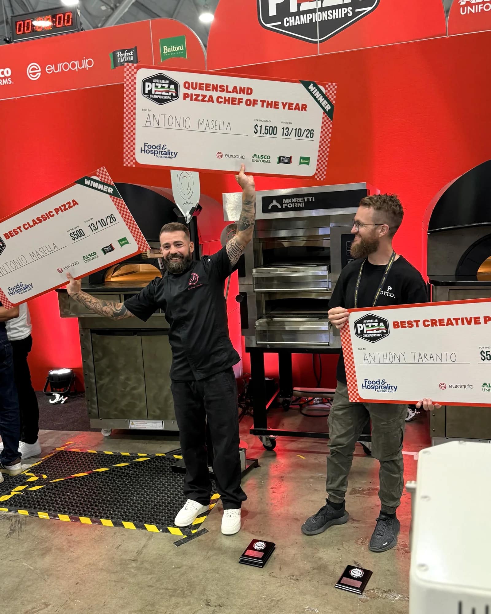 Antonio Masella raising his Queensland Pizza Chef of the Year and Best Classic Pizza cheques beside Anthony Taranto holding his Best Creative Pizza cheque, in front of pizza ovens on a red stage