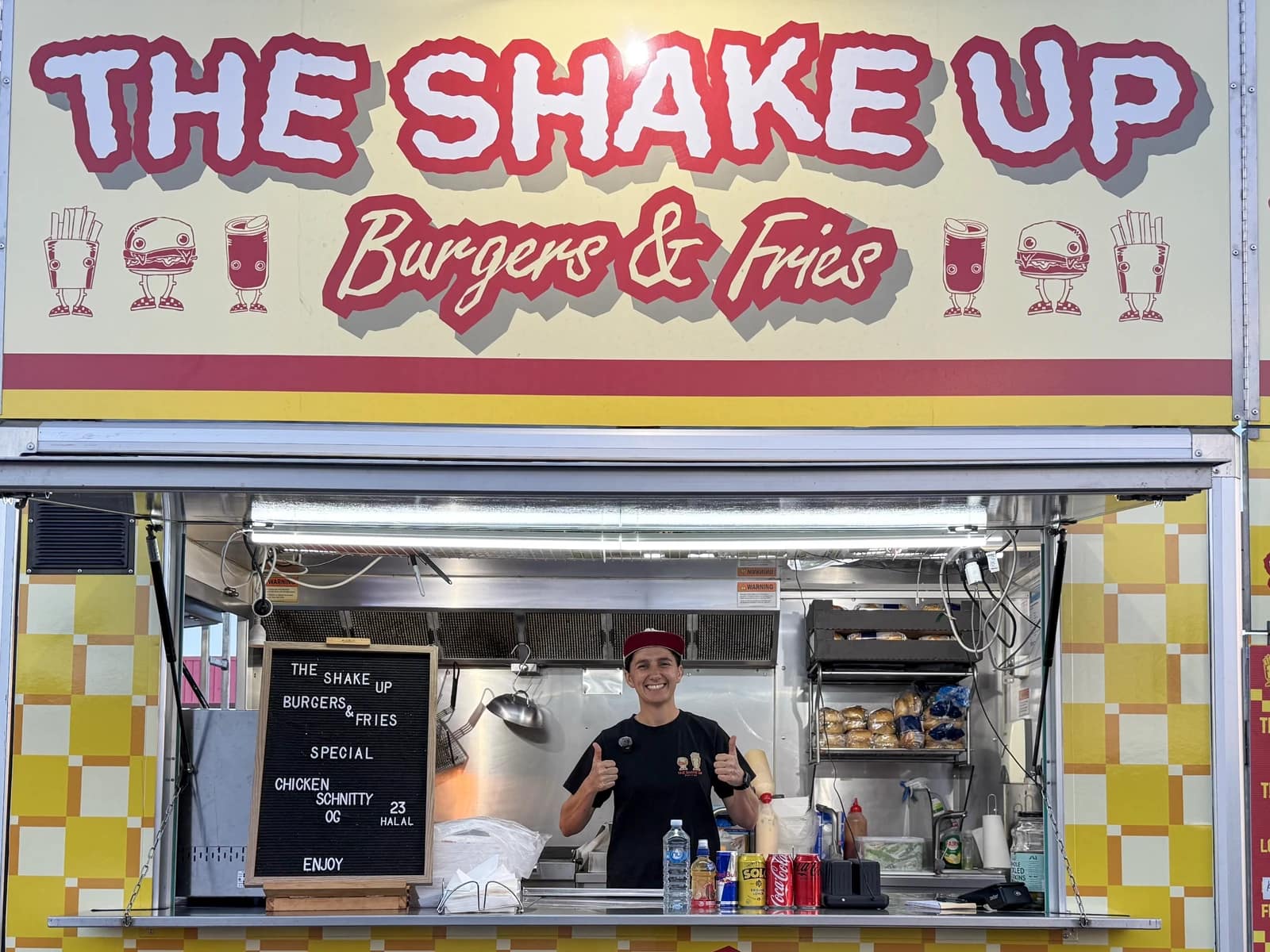 The Shake Up Burgers and Fries sign above its serving window, with Atiana smiling and giving two thumbs up.