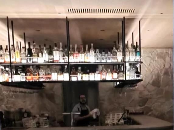 The Lodge bar seen through its glass door, with suspended bottle shelves above a stone wall and a bartender at work