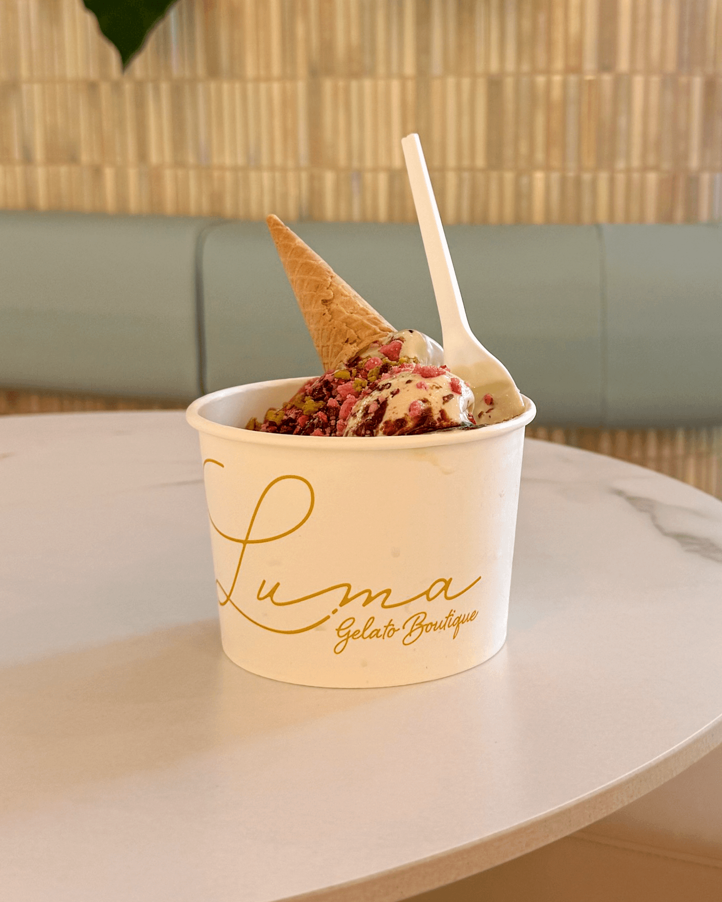 A Luma gelato cup with a cone and spoon on a table beside plants and mint seating