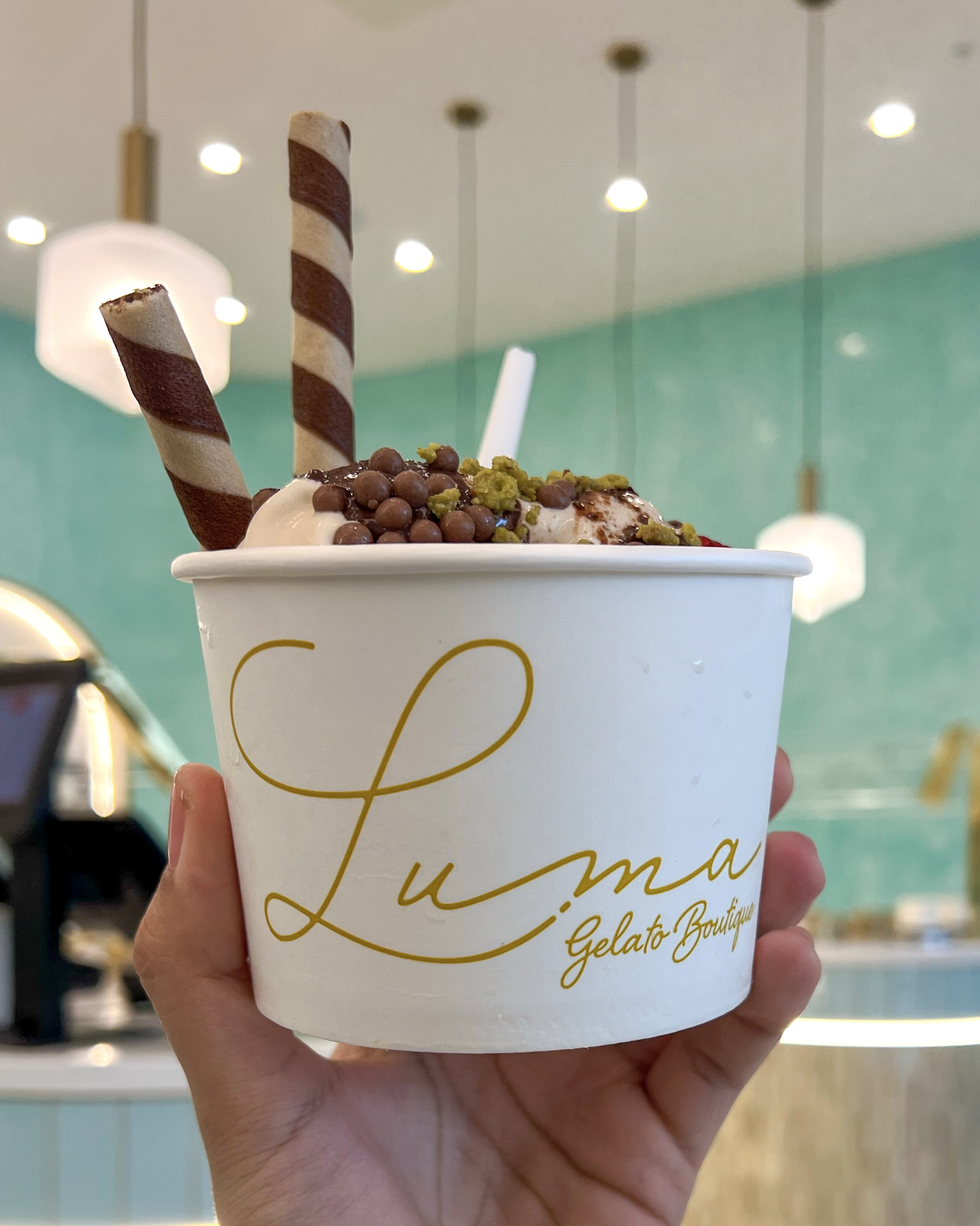 A hand holding a branded Luma gelato cup with toppings and wafer sticks inside the shop