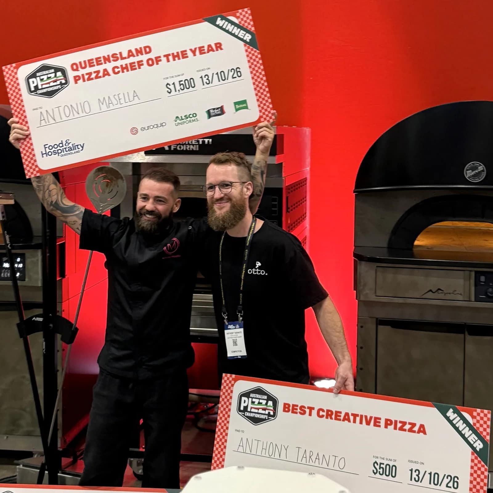 Antonio Masella and Anthony Taranto smiling together in front of pizza ovens, holding their winners' cheques