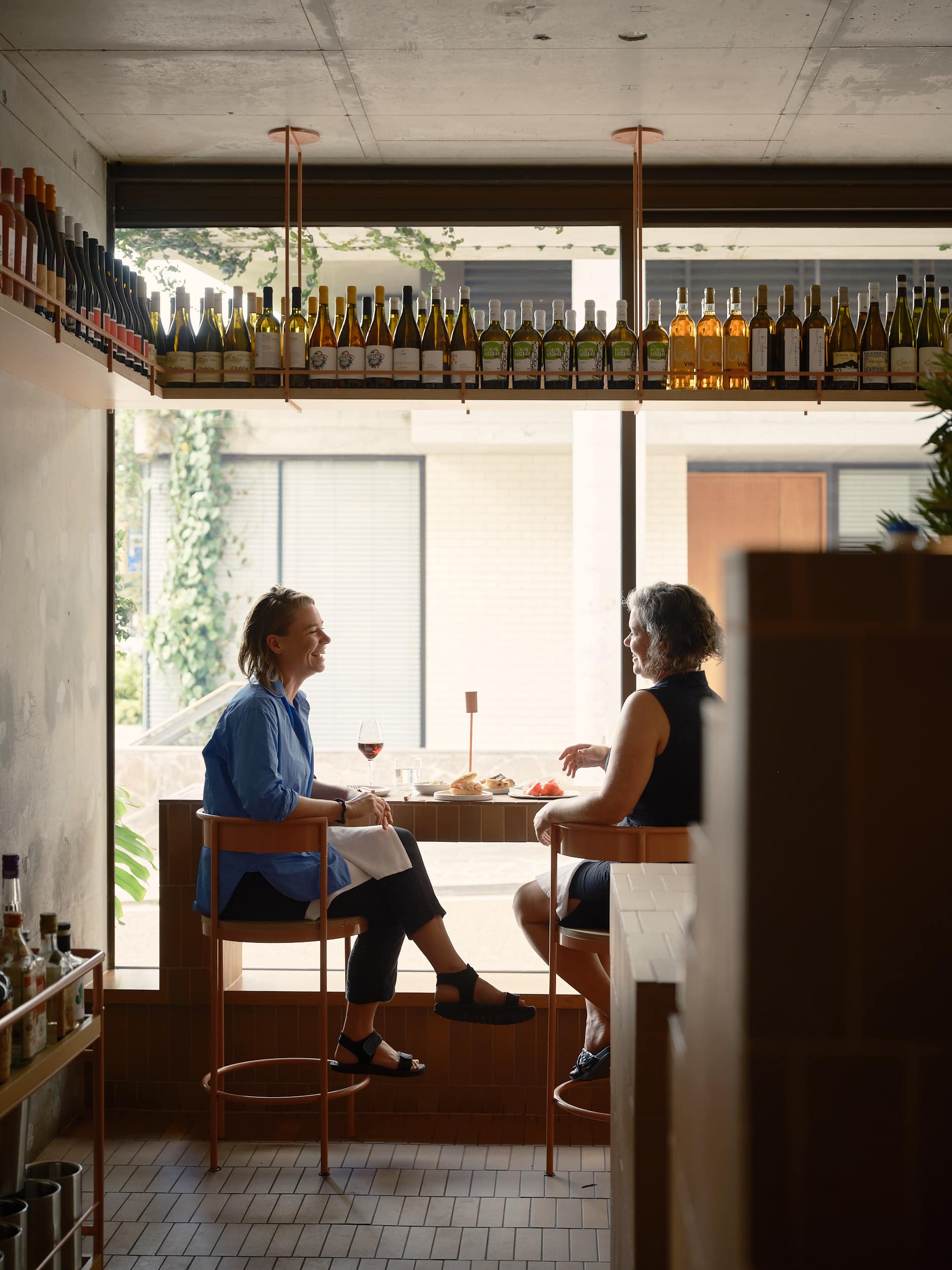 Two guests seated beside Biànca’s window beneath wine shelves